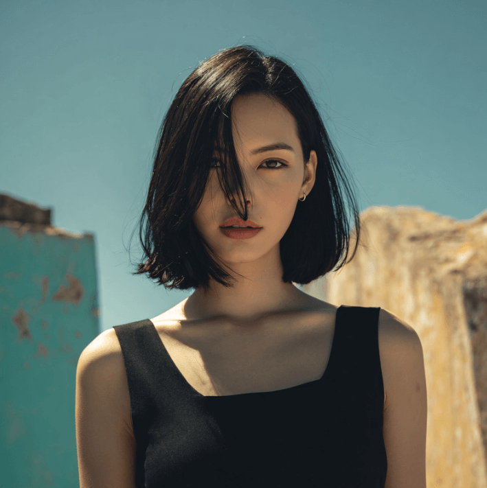 Portrait d'une femme avec un bob noir, des murs décolorés en arrière-plan sous un ciel bleu.