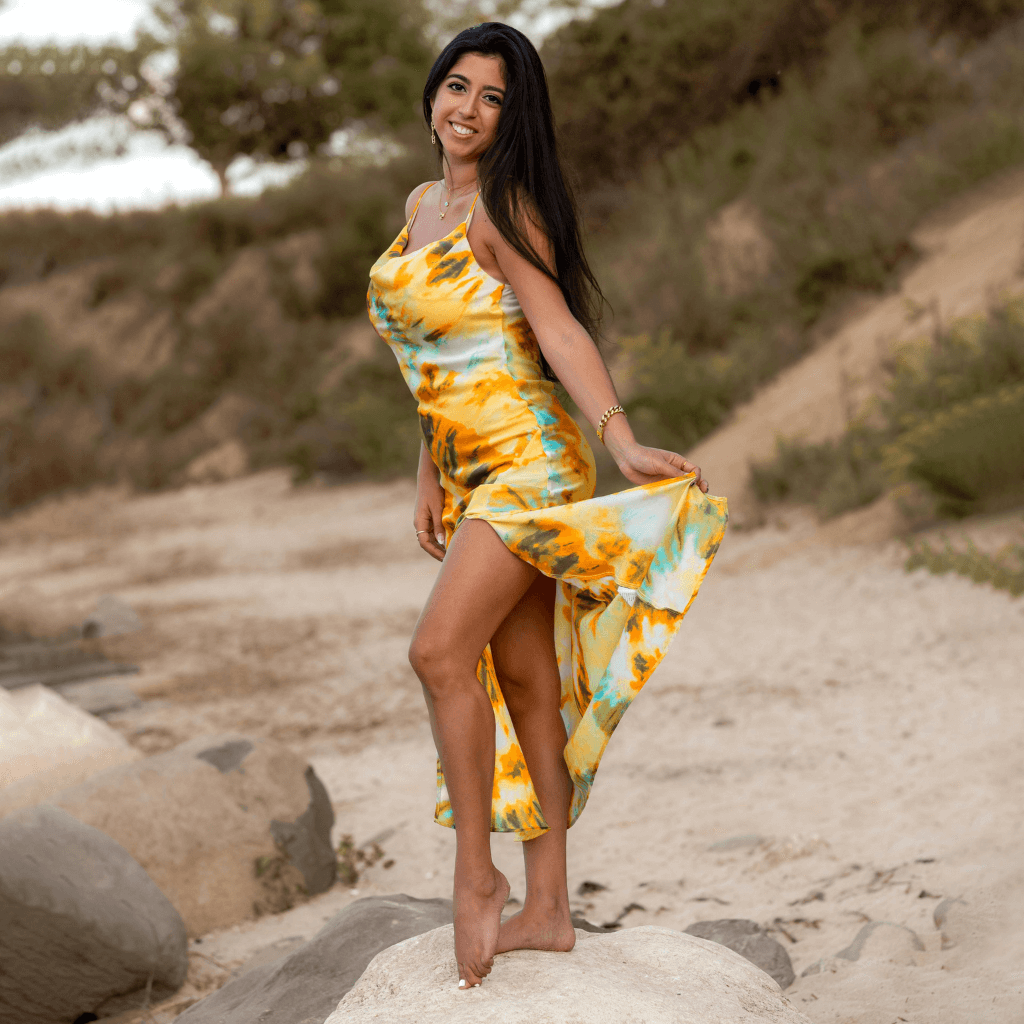 Femme souriante portant une robe colorée à motifs, marchant sur la plage avec un paysage naturel en arrière-plan.