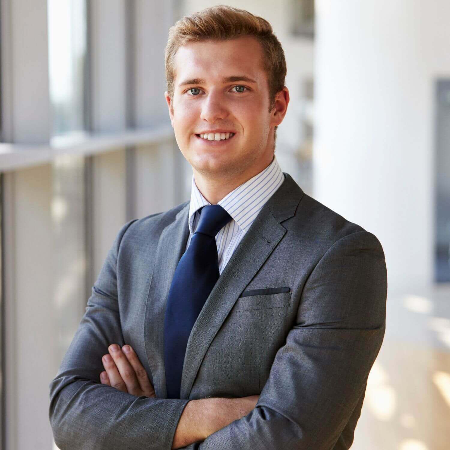 Homme d'affaires souriant en costume gris avec une cravate, debout dans un bureau moderne.